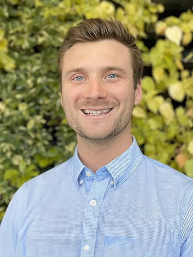 Smiling man with blue eyes wearing a light blue button-up shirt standing in front of leafy green background