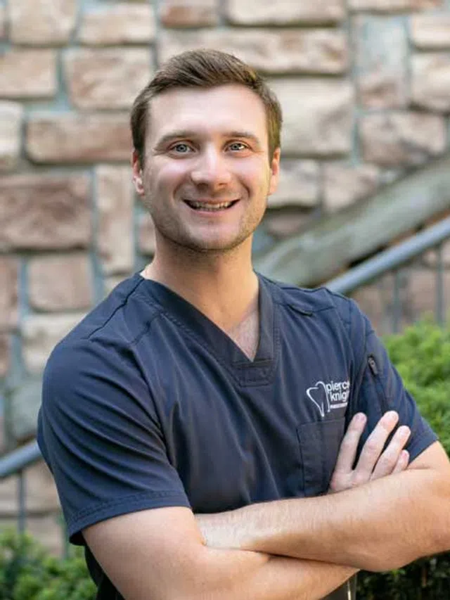 Smiling male healthcare professional in navy scrubs with crossed arms standing outdoors near stone wall