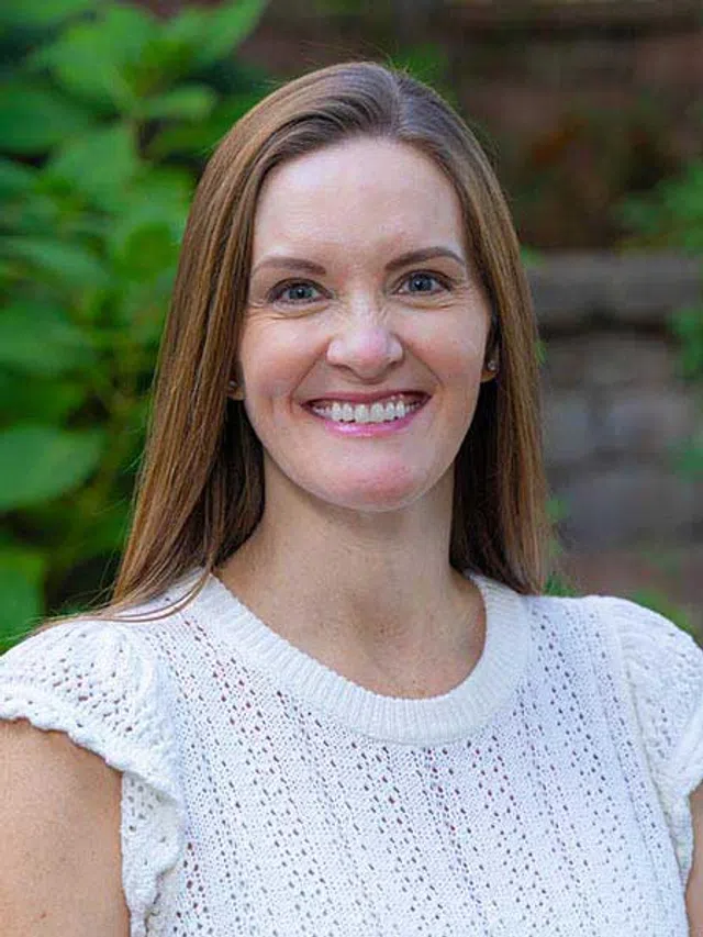 Smiling woman with long brown hair wearing a white knitted top standing outdoors with green foliage background