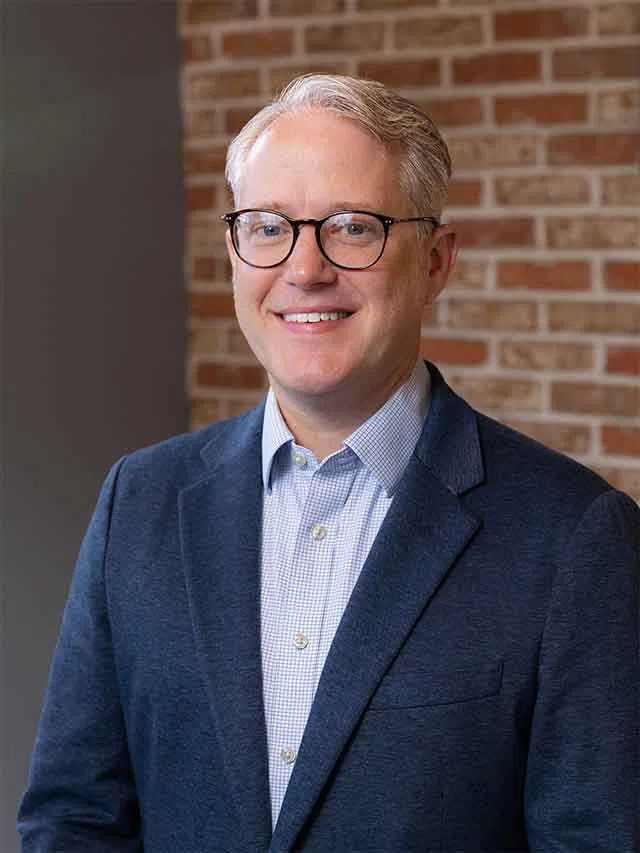 Smiling middle-aged man with glasses wearing a blue blazer and checkered shirt standing against a brick wall.
