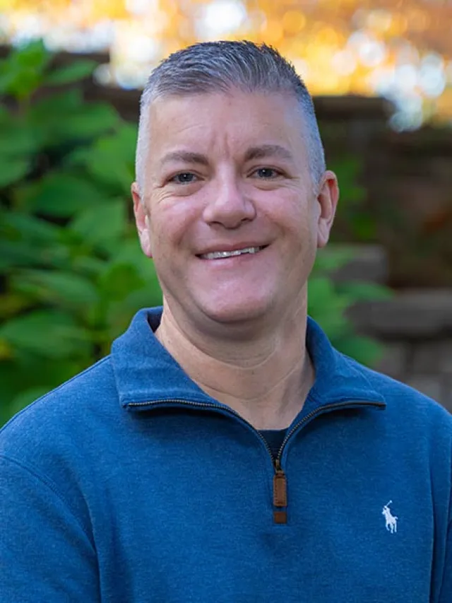 Smiling man wearing a blue quarter-zip sweater standing outdoors with green foliage and warm sunlight background.