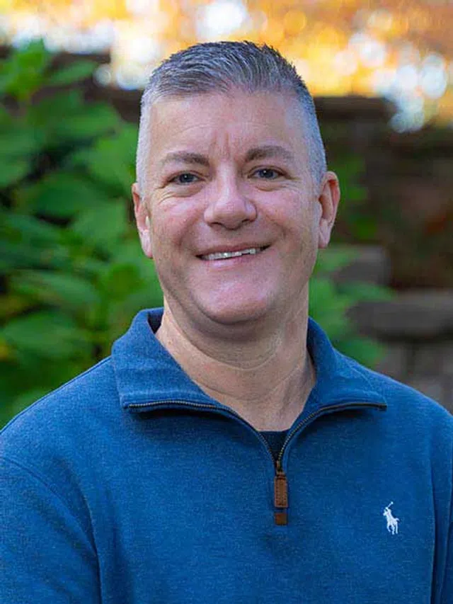 Smiling man wearing a blue quarter-zip sweater standing outdoors with greenery and soft sunlight in the background