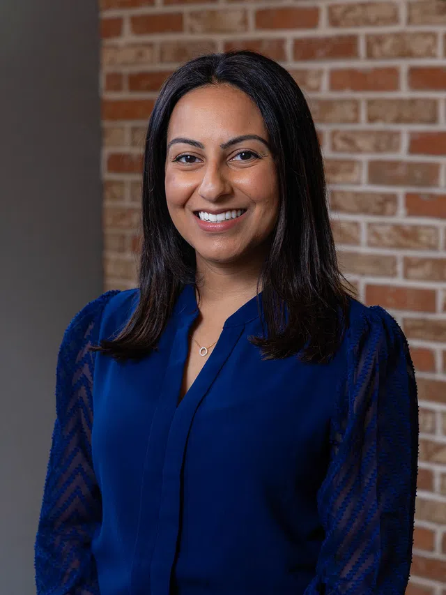 Smiling woman with long dark hair wearing a blue blouse standing in front of a brick wall background.
