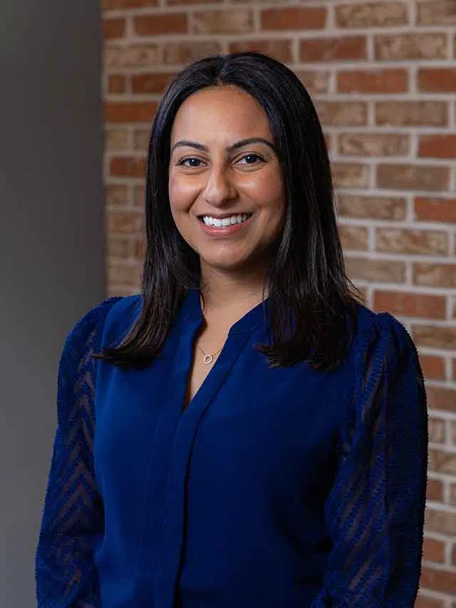 Smiling woman in blue blouse with long dark hair stands against a brick wall background