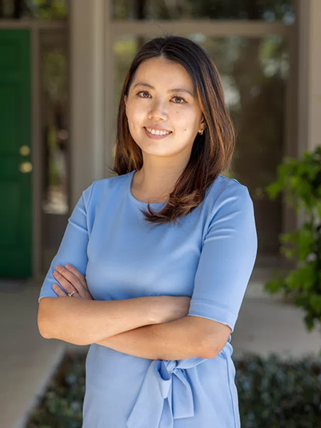 Smiling woman in light blue dress with crossed arms standing outdoors near green door and plants.