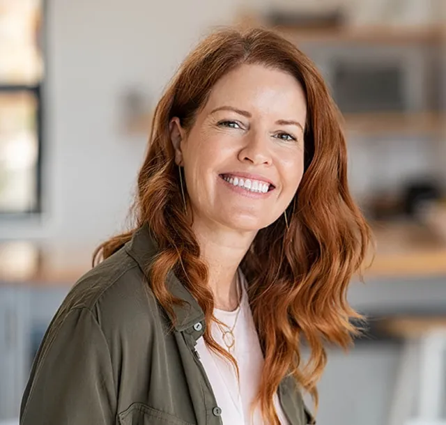 Smiling red-haired woman in casual outfit posing indoors with blurred background, radiating warmth and confidence