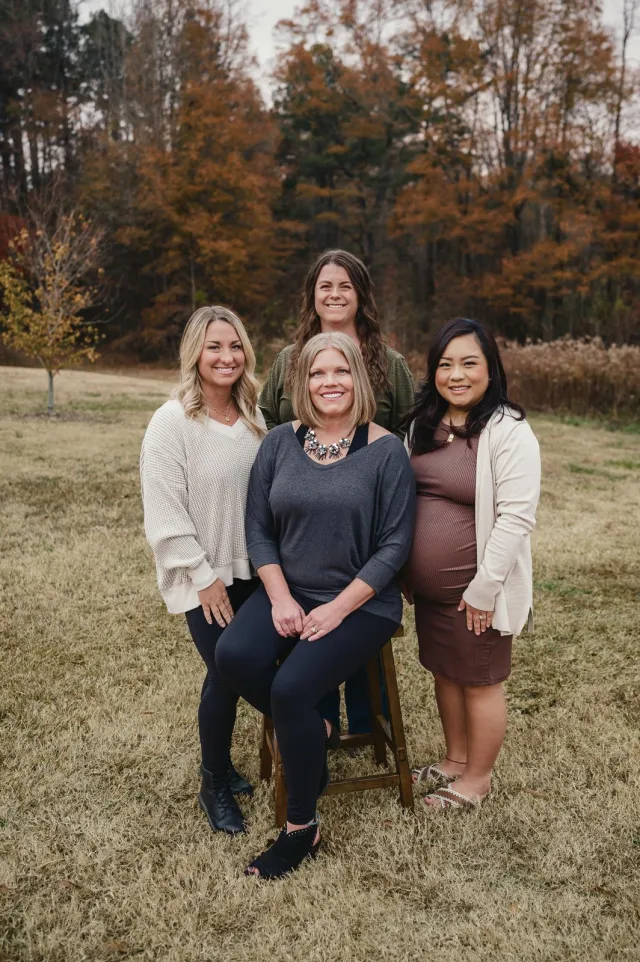 Group of four women posing outdoors in fall with autumn trees and brown grass in background