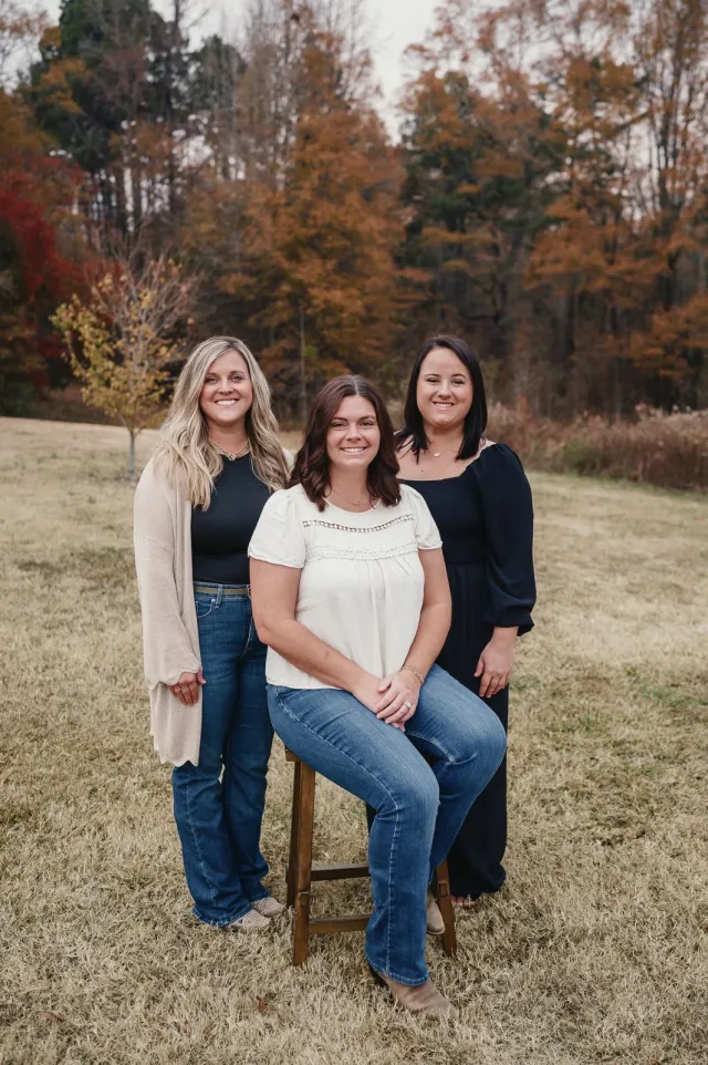 Three smiling women posing outdoors in autumn with trees and dry grass in the background.