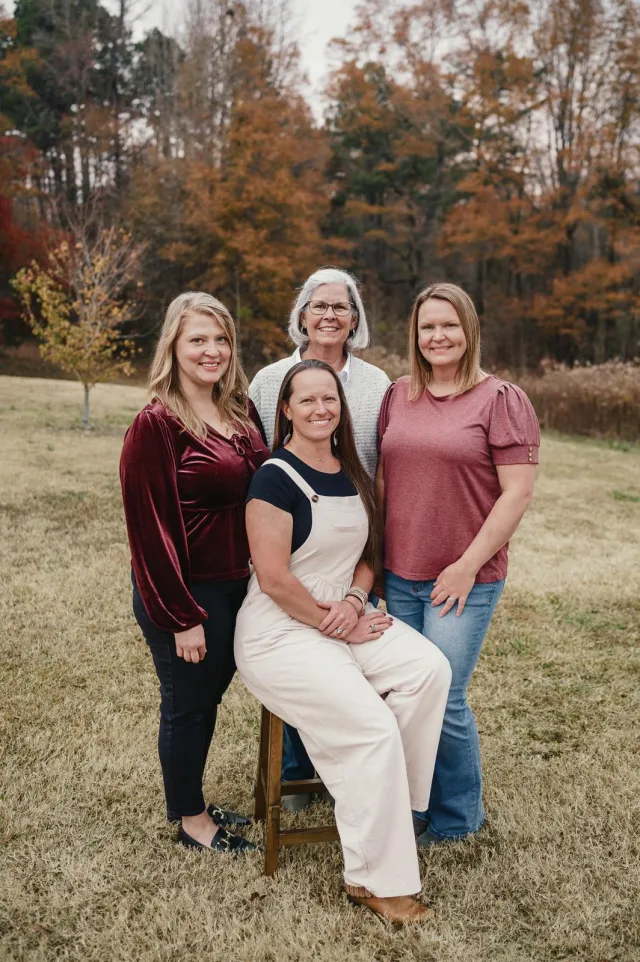 Four smiling women posing outdoors on autumn grass with fall-colored trees in the background