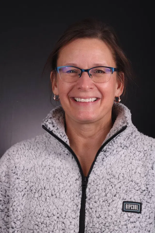 Smiling woman wearing blue glasses with a gray Rip Curl fleece jacket against a black background