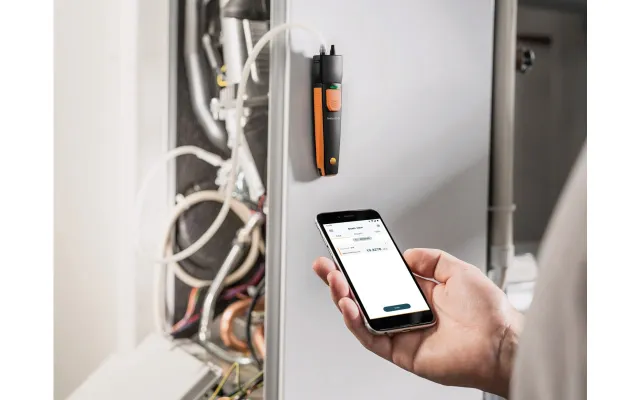 Hand holding smartphone displaying data, next to a black and orange device mounted on a gray panel with wires behind it.