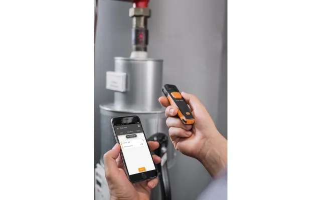 Person using smartphone and handheld device to scan a pipe sensor in an industrial setting.
