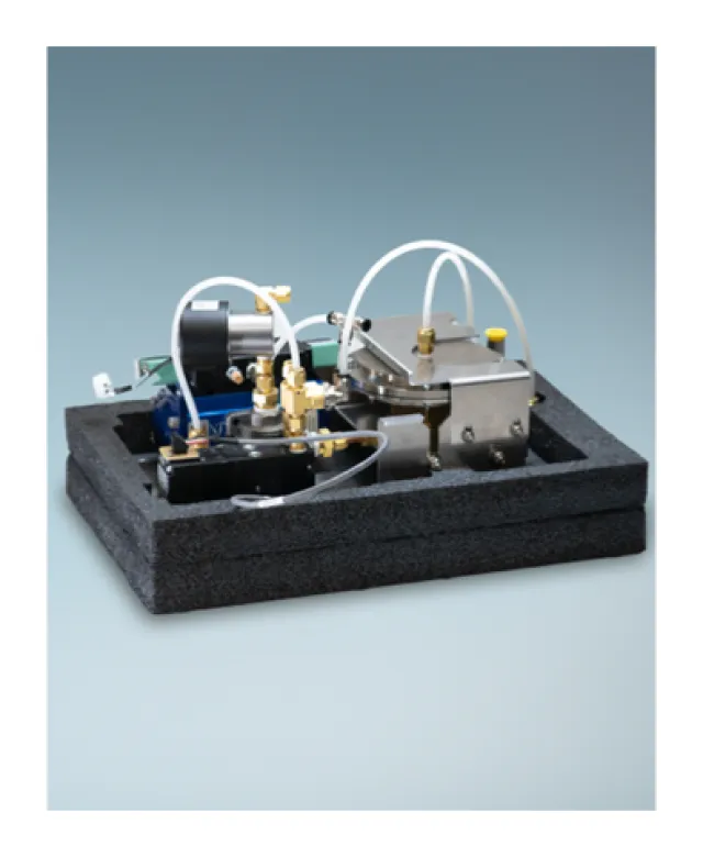 Compact mechanical device with metal components, tubes, and connectors mounted on black foam base against gray background.