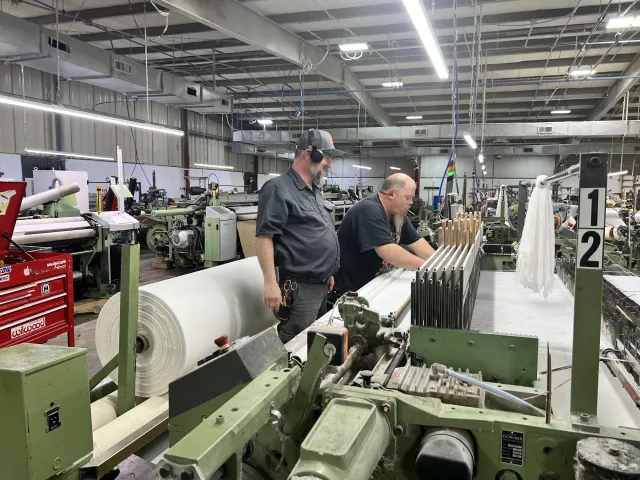 Two male workers operate large weaving machines producing fabric rolls in a brightly lit textile factory.