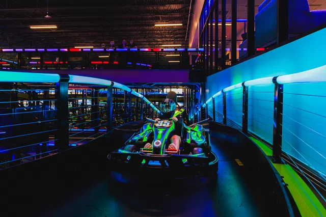Person driving a neon-lit go-kart on an indoor track with vibrant blue and purple lighting.