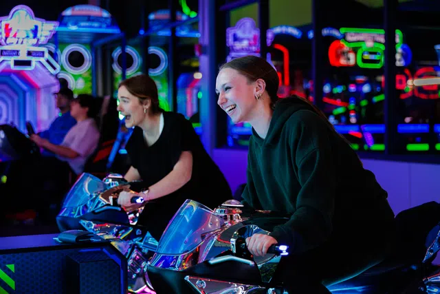 Two young women enjoying a lively arcade game riding motorcycle simulators amid colorful neon lights.