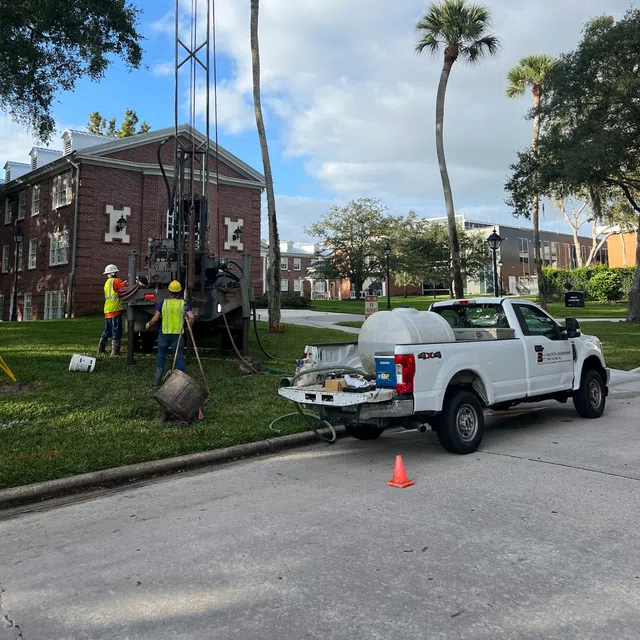 Workers in safety vests operating drilling equipment near a white utility truck on a residential street with palm trees.