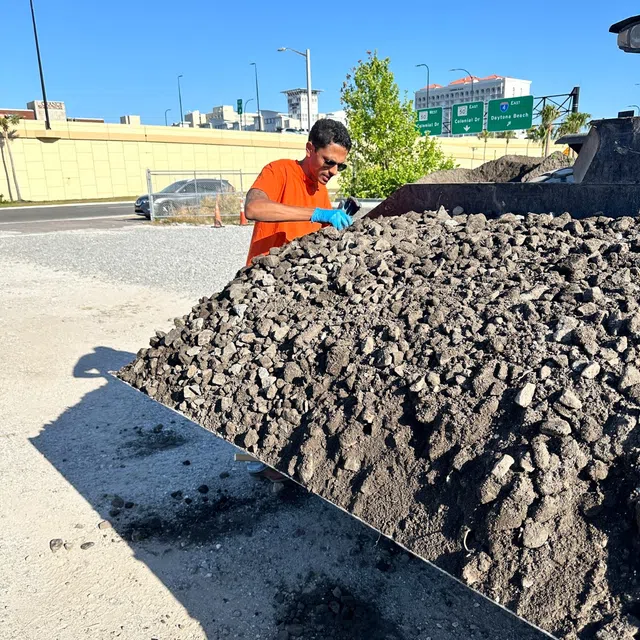 Worker in orange shirt examines large pile of gravel or crushed stone outdoors on sunny day.