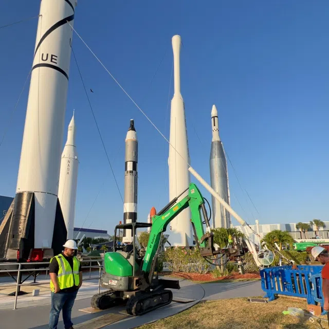 Workers with a green excavator in front of tall white rockets displayed outdoors under a clear blue sky at NASA.