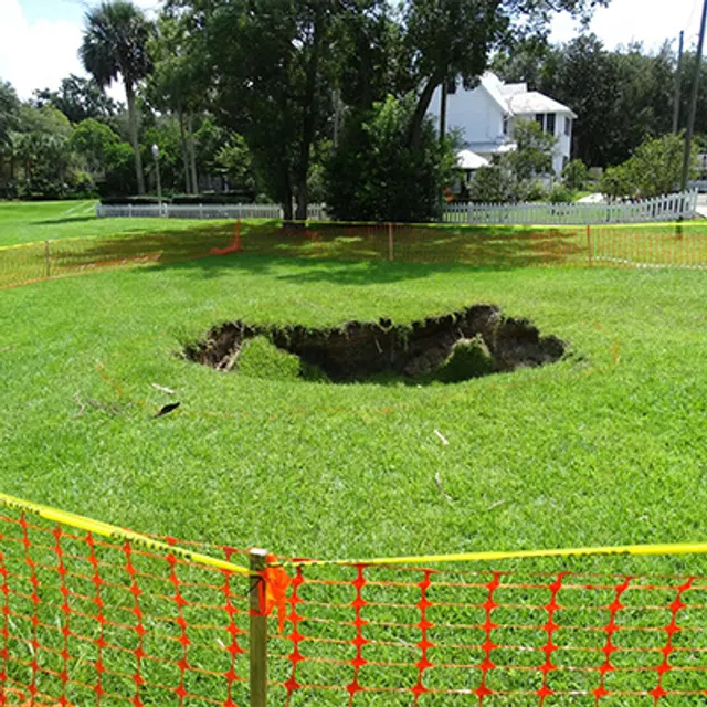 Large sinkhole in green grass fenced off with orange plastic safety barrier near residential area