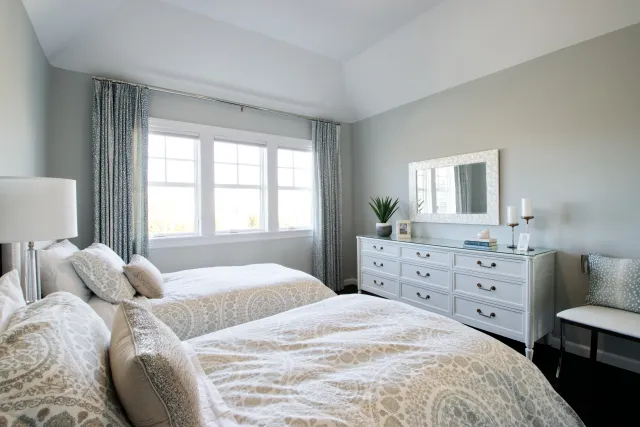 Bright bedroom with two twin beds, patterned bedding, large window, dresser with mirror, and soft gray walls.