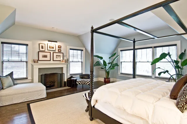 Bright bedroom with four-poster bed, fireplace, seating area, potted plants, and large windows with blinds