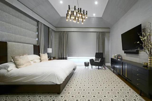 Modern bedroom with king-size bed, patterned carpet, chandelier, black armchair, and large wall-mounted TV