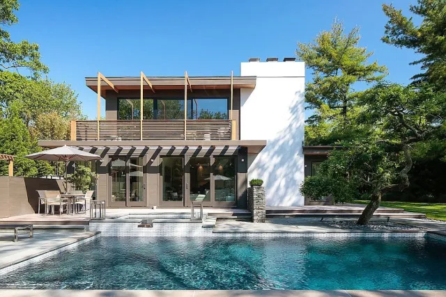 Modern two-story house with large windows, outdoor dining area, and a clear blue swimming pool surrounded by trees.