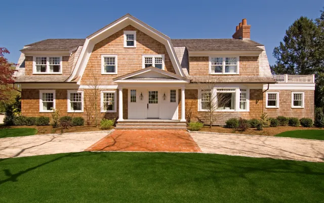Large two-story house with cedar shingles, white trim, green lawn, and brick walkway under clear blue sky.