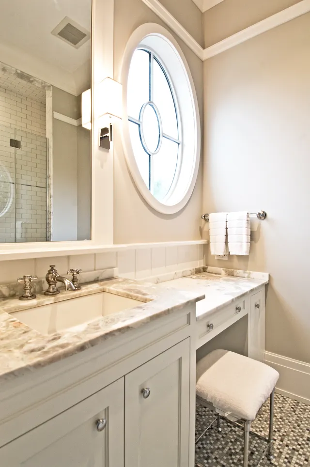 Bright modern bathroom with marble countertop, round window, white vanity, and tiled floor under soft lighting.