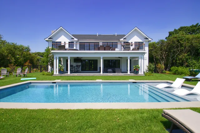 Modern two-story white house with large pool, green lawn, outdoor seating, and clear blue sky