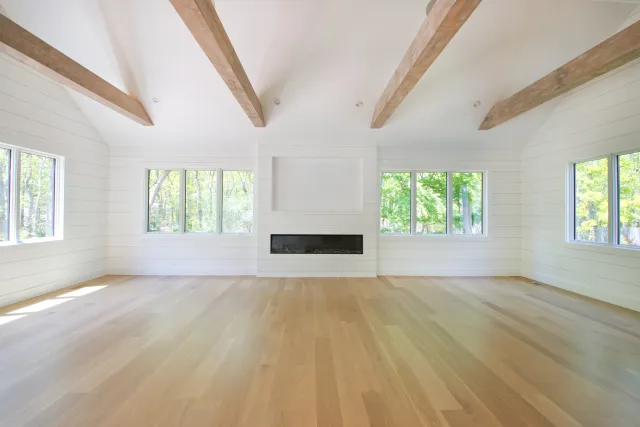 Bright empty room with hardwood floors, white walls, wooden ceiling beams, large windows, and a modern built-in fireplace.
