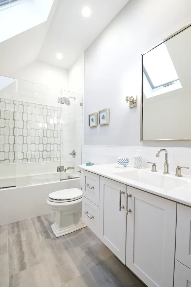 Bright modern white bathroom with glass shower, toilet, vanity, large mirror, and skylight window.