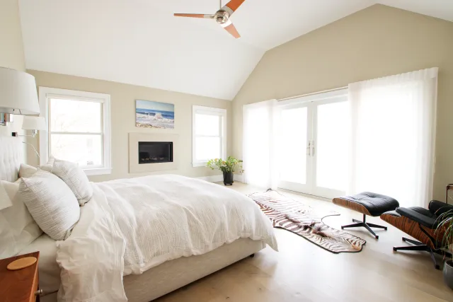 Bright modern bedroom with white bedding, vaulted ceiling, ceiling fan, fireplace, and zebra rug near lounge chair.