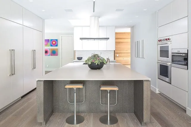 Modern white kitchen with large island, two bar stools, wood flooring, and colorful wall art in background