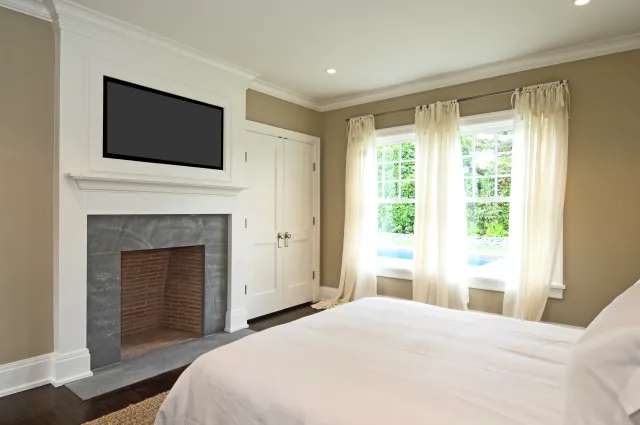 Bright bedroom with white bedding, large window with sheer curtains, fireplace, and wall-mounted TV.
