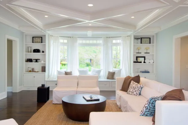 Bright living room with white sofas, patterned pillows, round wooden coffee table, and large window with sheer curtains