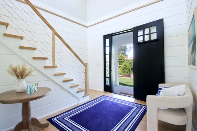 Bright modern entryway with black door, wooden staircase, round table, blue rug, and cozy seating area.