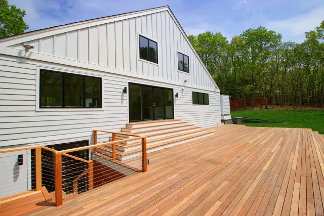 Modern white house with large wooden deck and stairs surrounded by green forest under blue sky.