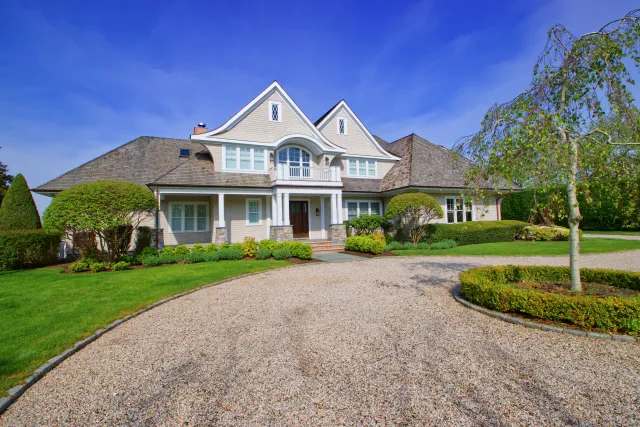 Large modern suburban house with manicured lawn, gravel driveway, and clear blue sky on a sunny day