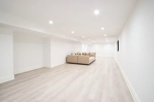Bright spacious basement with light wood flooring, recessed lighting, and a beige sectional sofa with pillows.