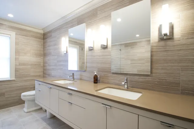 Modern bathroom with double sinks, large mirrors, beige countertop, wall-mounted lights, and wooden tile walls.