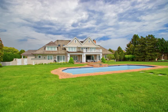 Large white house with a sloped roof and balcony overlooking a rectangular pool with brick edging on a green lawn.