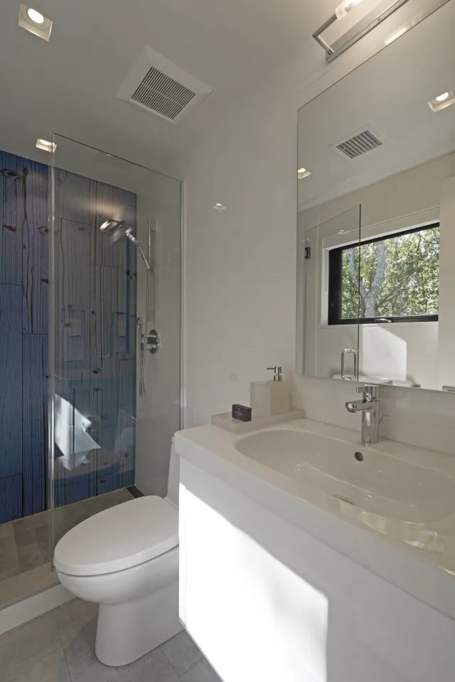Bright modern bathroom with white fixtures, glass shower enclosure with blue accent wall, and large mirror