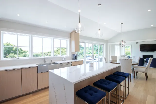 Bright modern kitchen with marble island, navy blue stools, wooden cabinets, large windows, and dining area.