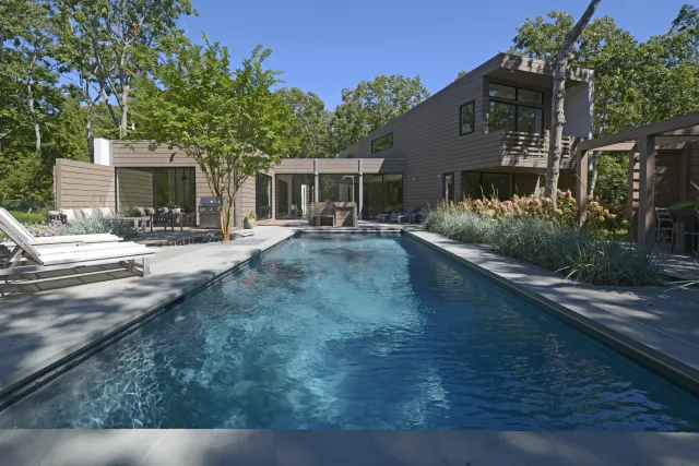Modern house with a rectangular swimming pool, lounge chairs, and surrounding trees under a clear sky.