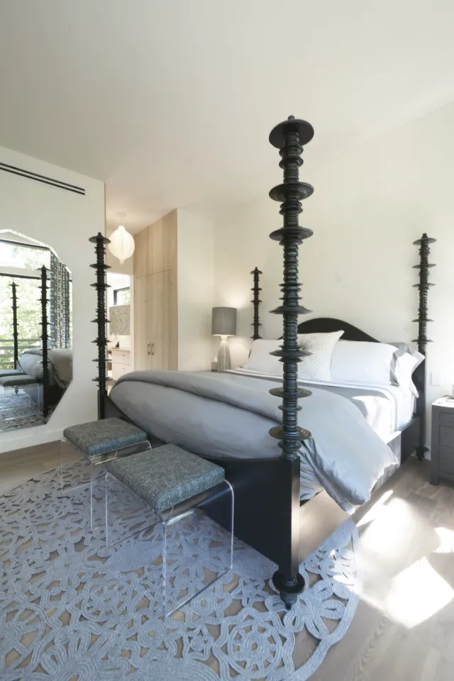 Bright bedroom with a black four-poster bed, gray bedding, two stools, and a large decorative rug.