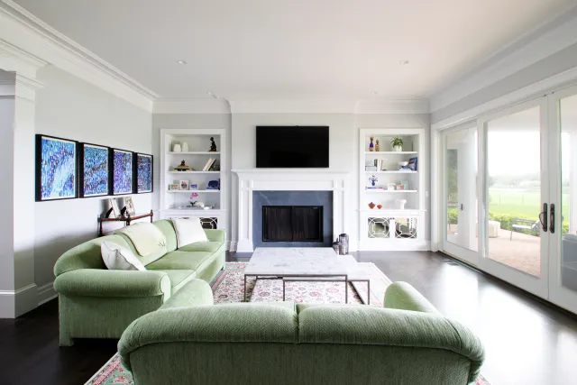 Bright living room with green sofas, fireplace, wall-mounted TV, built-in shelves, and large glass doors to patio.