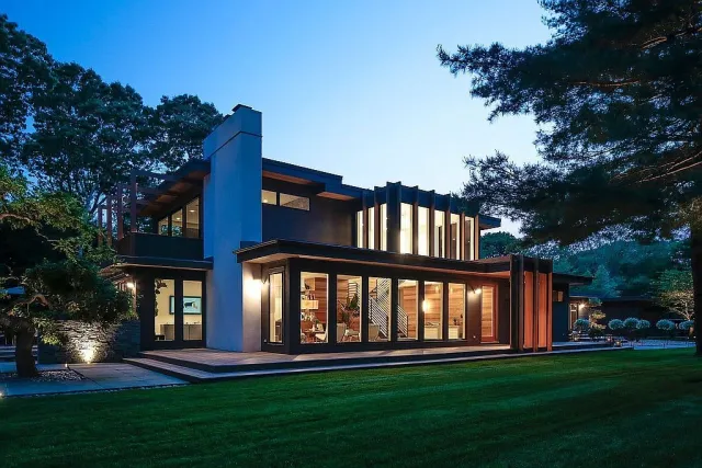 Modern two-story house with large windows and outdoor lighting set in a green landscaped yard at dusk.