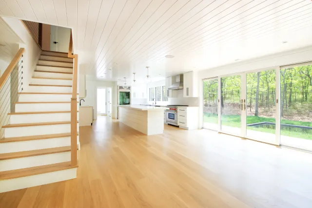 Bright modern open-concept kitchen with wooden floors, white cabinetry, staircase, and large sliding glass doors to forest.